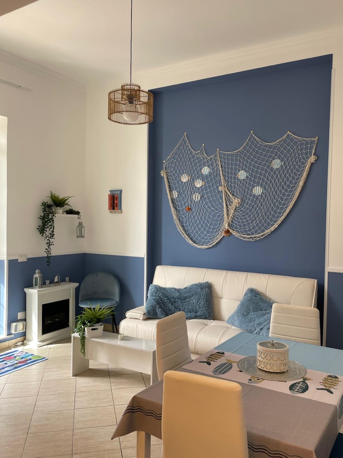 A light-filled living area features a white sofa adorned with blue cushions, complemented by a decorative fishing net on the navy blue wall. A small dining table sits nearby, adding to the functional layout, while plants introduce a touch of greenery to the space.