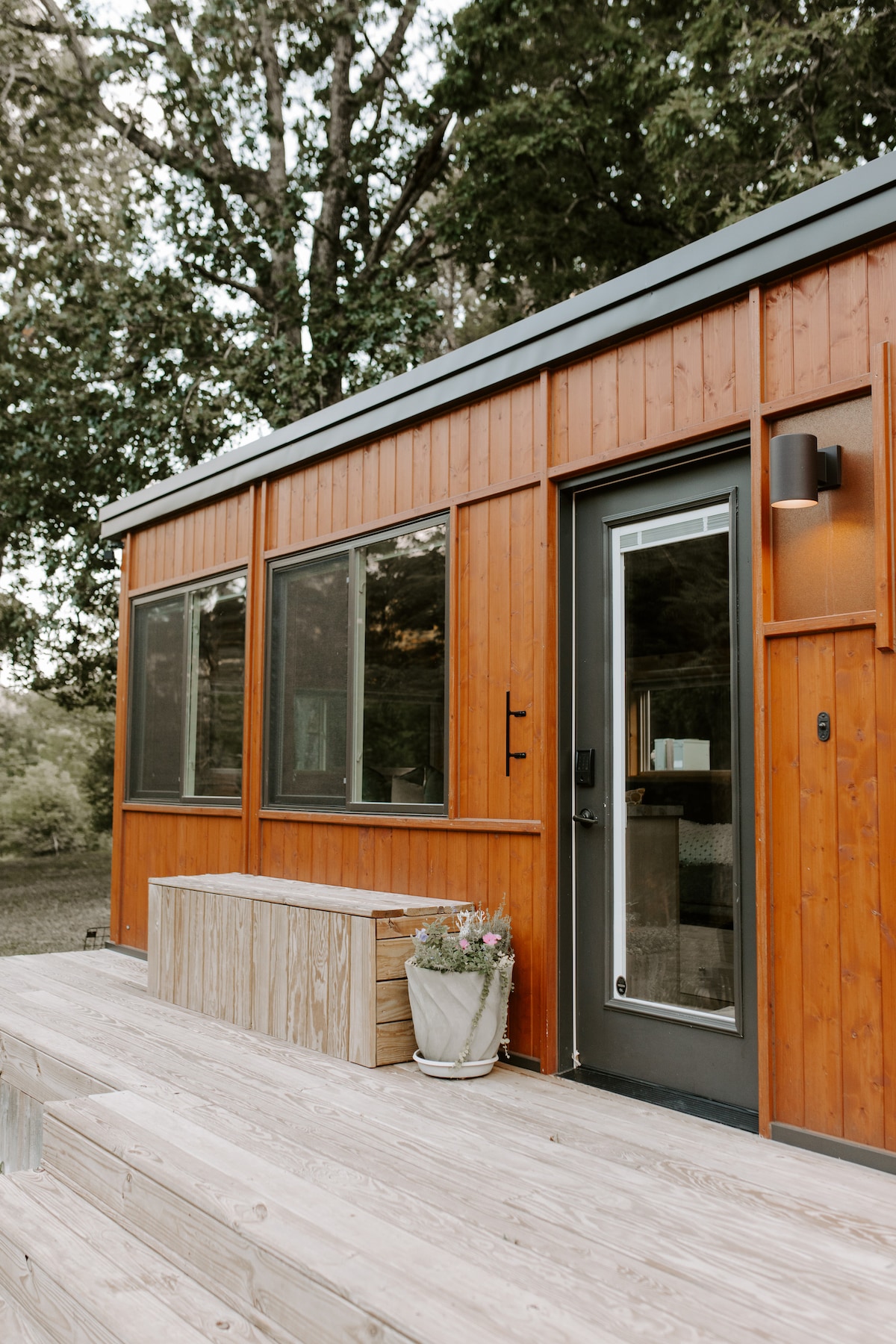The exterior of the tiny home features warm wooden siding and multiple large windows, providing a view of the surrounding nature. A simple wooden step leads to the entrance, where a potted plant adds a touch of greenery beside the door.
