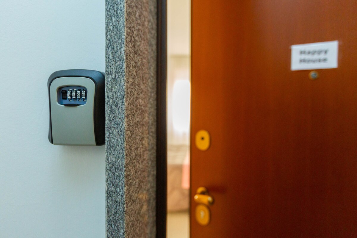 A wall-mounted safe with a numeric combination lock is positioned beside a wooden door. The door features a circular knob and a small sign labeled 'Happy Home.' Soft light is visible in the background, indicating a well-lit interior space.