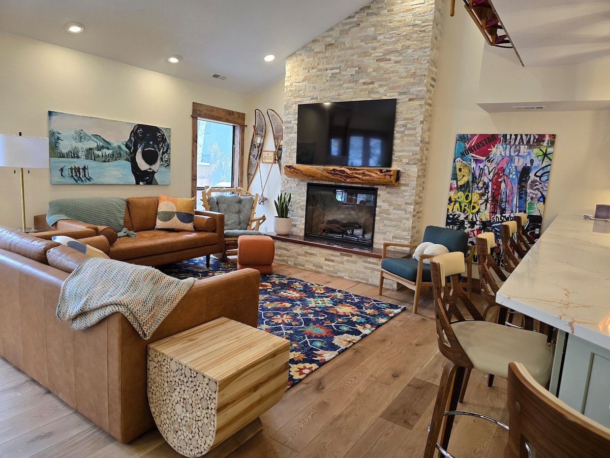 A spacious living area features a large brown leather sofa, complemented by colorful artwork and vibrant decor. A stone-surrounded fireplace stands beneath a wall-mounted television. Natural light enters through large windows, illuminating a patterned area rug and an inviting seating arrangement.