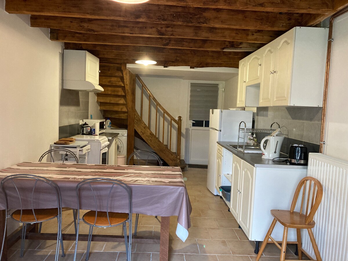 A spacious kitchen and living area is present, featuring wooden beams overhead. A dining table with metal chairs sits in the foreground, while a staircase leads to the upper level. Kitchen appliances include a refrigerator, stove, and microwave, all arranged along the walls.