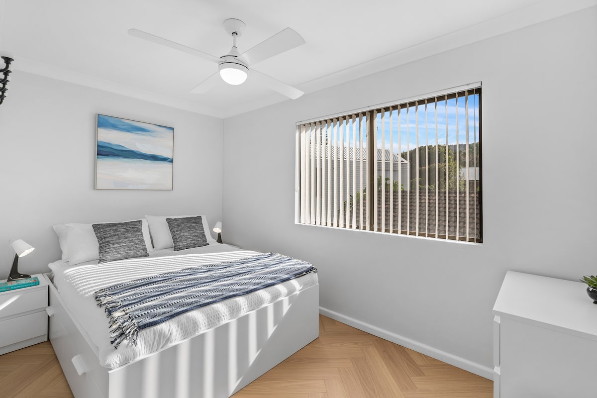 The second bedroom features a queen bed dressed in soft textures, with sunlight streaming through wide windows and peaceful artwork completing the calm vibe.