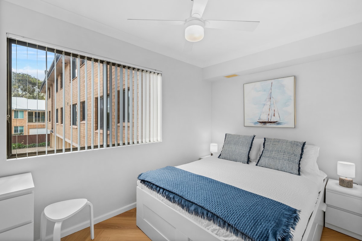 Relax in the primary bedroom where a queen bed wrapped in crisp white linens, ocean-toned details, and a ceiling fan create a restful, breezy retreat at the end of each day.