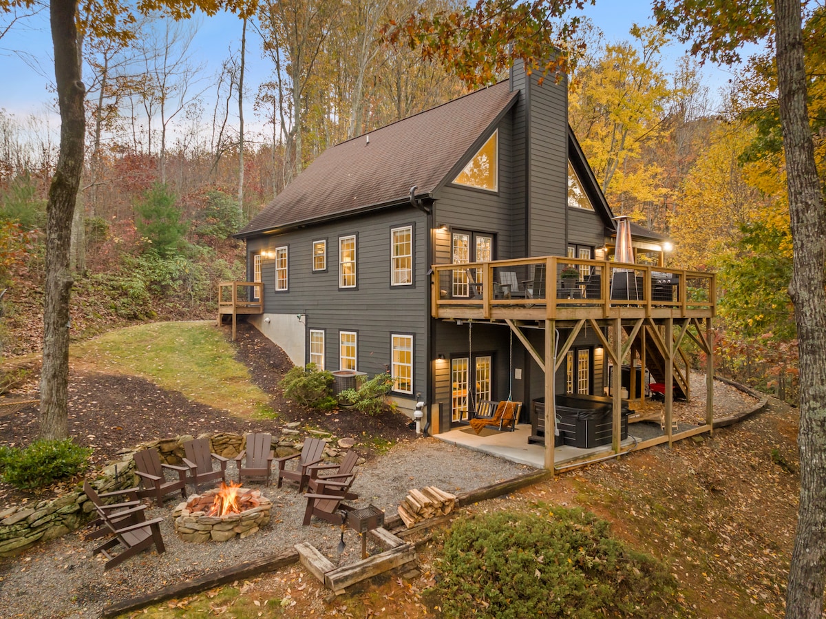 The exterior view of the modern cabin showcases multiple levels with large windows. A spacious deck overlooks the surrounding wooded area, while a firepit area is nestled in the yard, featuring ample seating around a stone circle. Autumn foliage enhances the rustic charm.