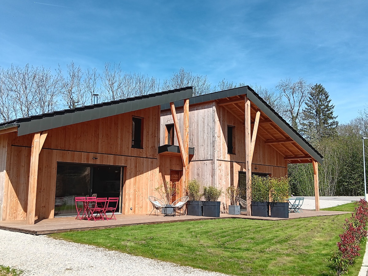 The modern wooden lodge features a spacious deck with outdoor seating, surrounded by well-maintained greenery. Large glass doors allow natural light into the interior, while the sloped roof and natural materials create a harmonious connection with the surrounding landscape.