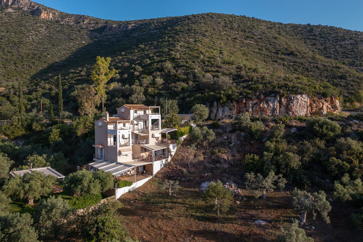 A villa is positioned on a hillside, surrounded by greenery and rocky terrain. The multi-level structure features large balconies and terraces that overlook the scenic landscape, providing a sense of harmony with nature.