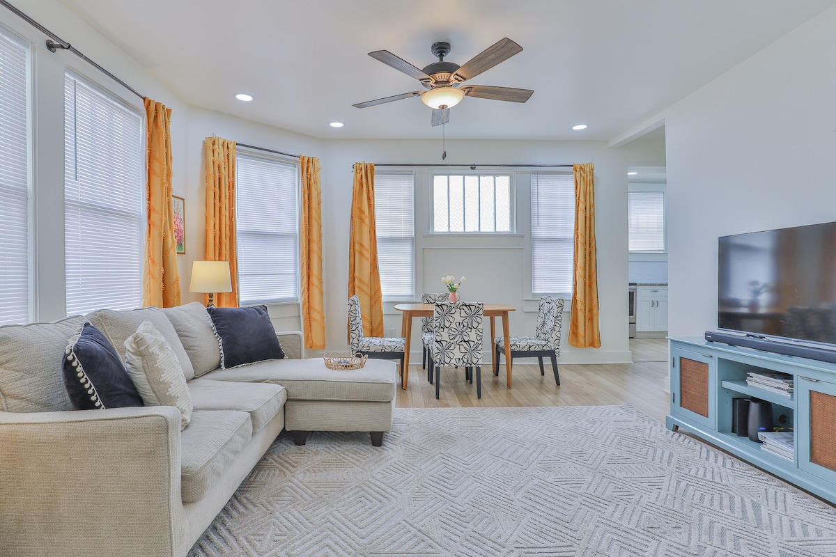 A light-filled living area features a comfortable sectional sofa accompanied by decorative cushions. A wooden dining table is set for two, surrounded by patterned chairs. Large windows draped with orange curtains allow natural light to enhance the space, while a flat-screen TV is positioned for easy viewing.