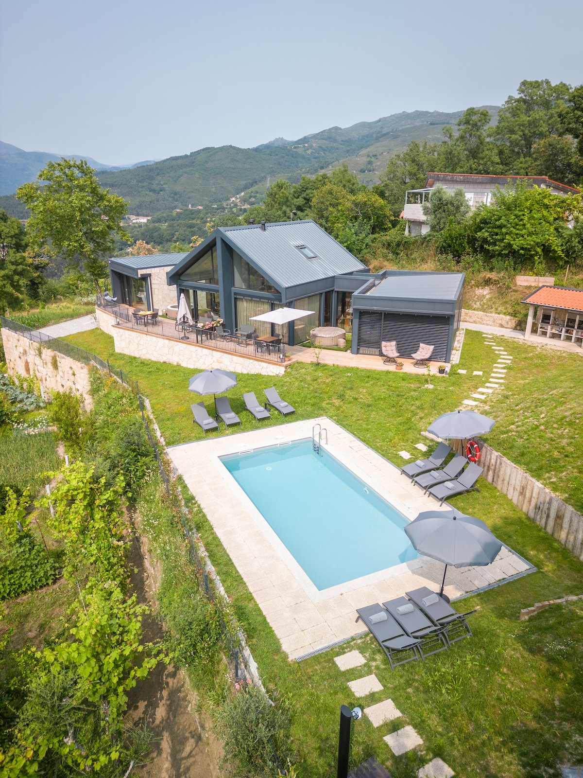An aerial view of the property reveals a modern home with large windows, surrounded by lush greenery. A saltwater pool is positioned in the backyard, accompanied by lounge chairs and umbrellas. A terraced area showcases a spacious patio for outdoor relaxation.