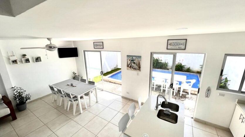 An open-concept living area showcases a dining table surrounded by multiple chairs, and a kitchen counter with modern faucets. Large windows provide a view of the private pool outside. Neutral-colored walls and tiled floors contribute to a spacious and bright environment.