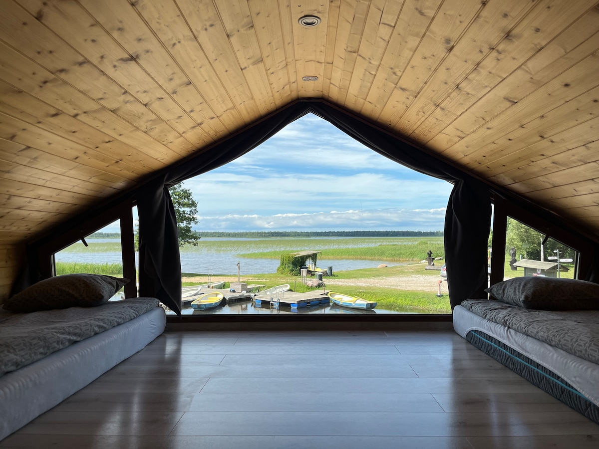 The interior of a loft area is showcased, featuring two beds positioned beside large windows. The view displays serene lake waters and lush greenery. Wooden beams and a sloped ceiling add a warm touch, while natural light brightens the space.