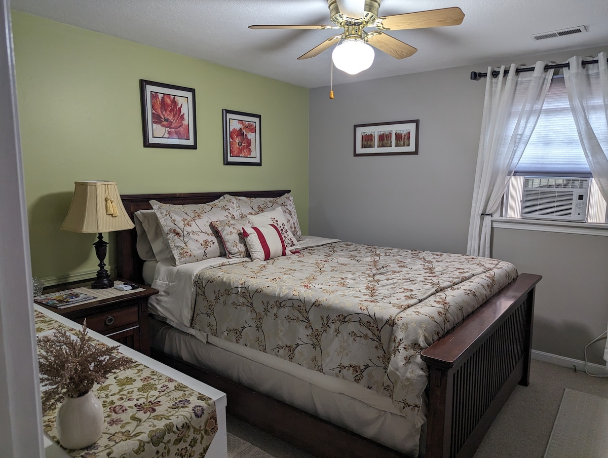 A cozy bedroom features a spacious bed adorned with a floral comforter and accented by decorative pillows. Soft light is provided by a ceiling fan. Two framed wall art pieces complement the light green walls, while a nightstand and dresser complete the inviting space.