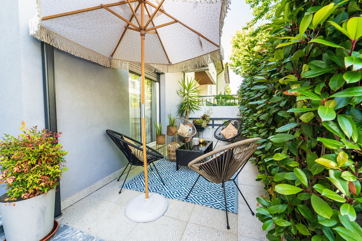 A shaded patio area is presented with a large umbrella and two comfortable chairs. A decorative rug lies on the floor, while potted plants and natural greenery provide a serene backdrop, creating a peaceful outdoor retreat for relaxation.