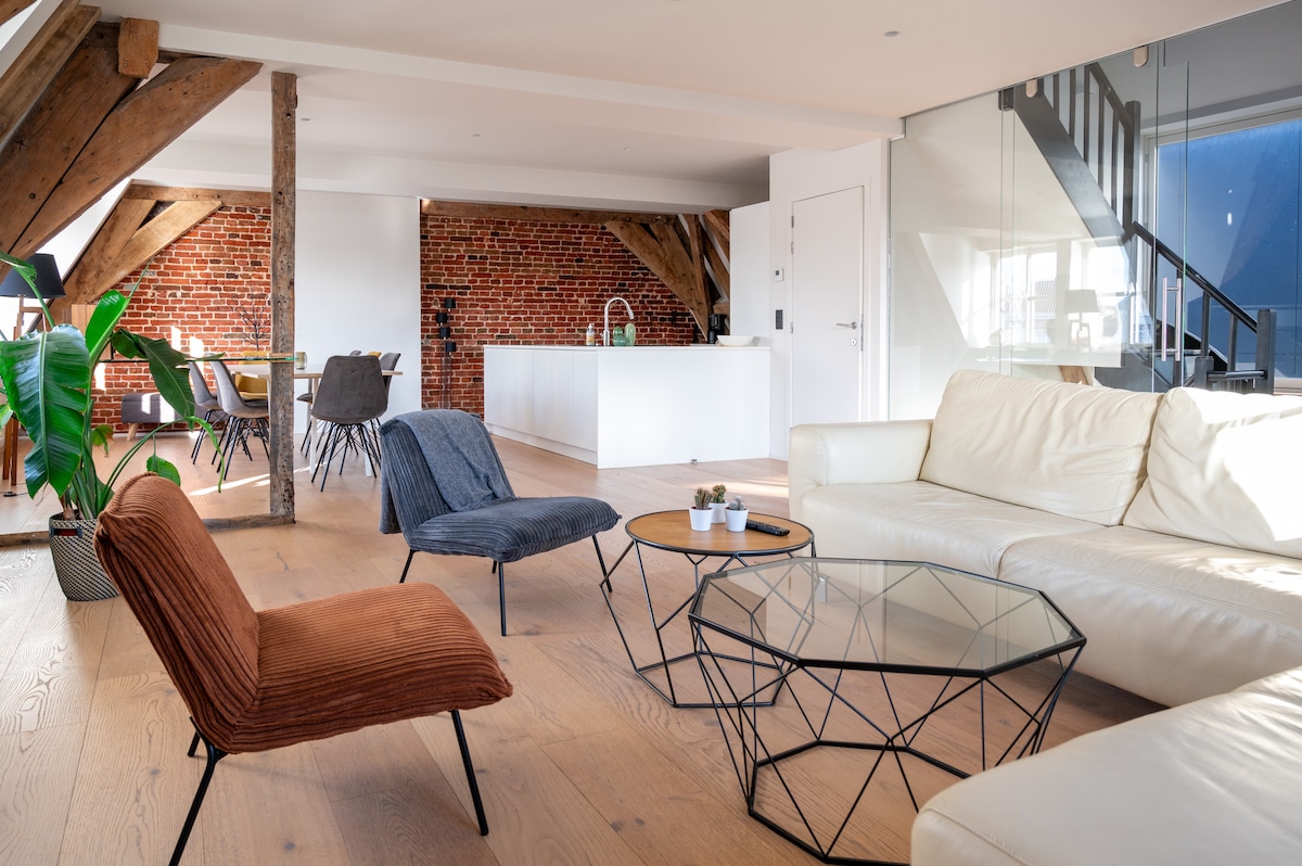 The spacious living area features a mix of comfortable seating, including an orange chair and two textured blue chairs, arranged around a glass coffee table. Exposed wooden beams and a brick wall add character, while a modern kitchen is visible in the background.