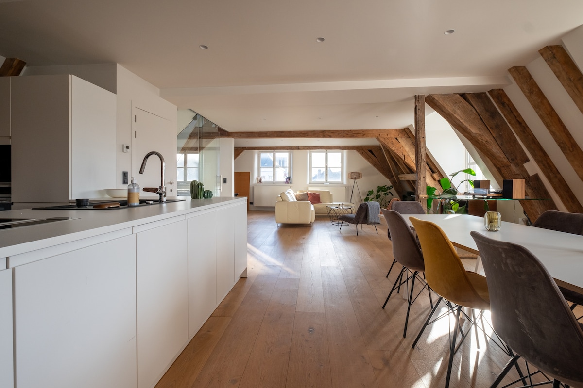 The spacious living area features modern furnishings with a fully equipped kitchen and a large dining table. Exposed wooden beams add character to the ceiling. Natural light fills the room through multiple windows, enhancing the bright and airy feel of the space.