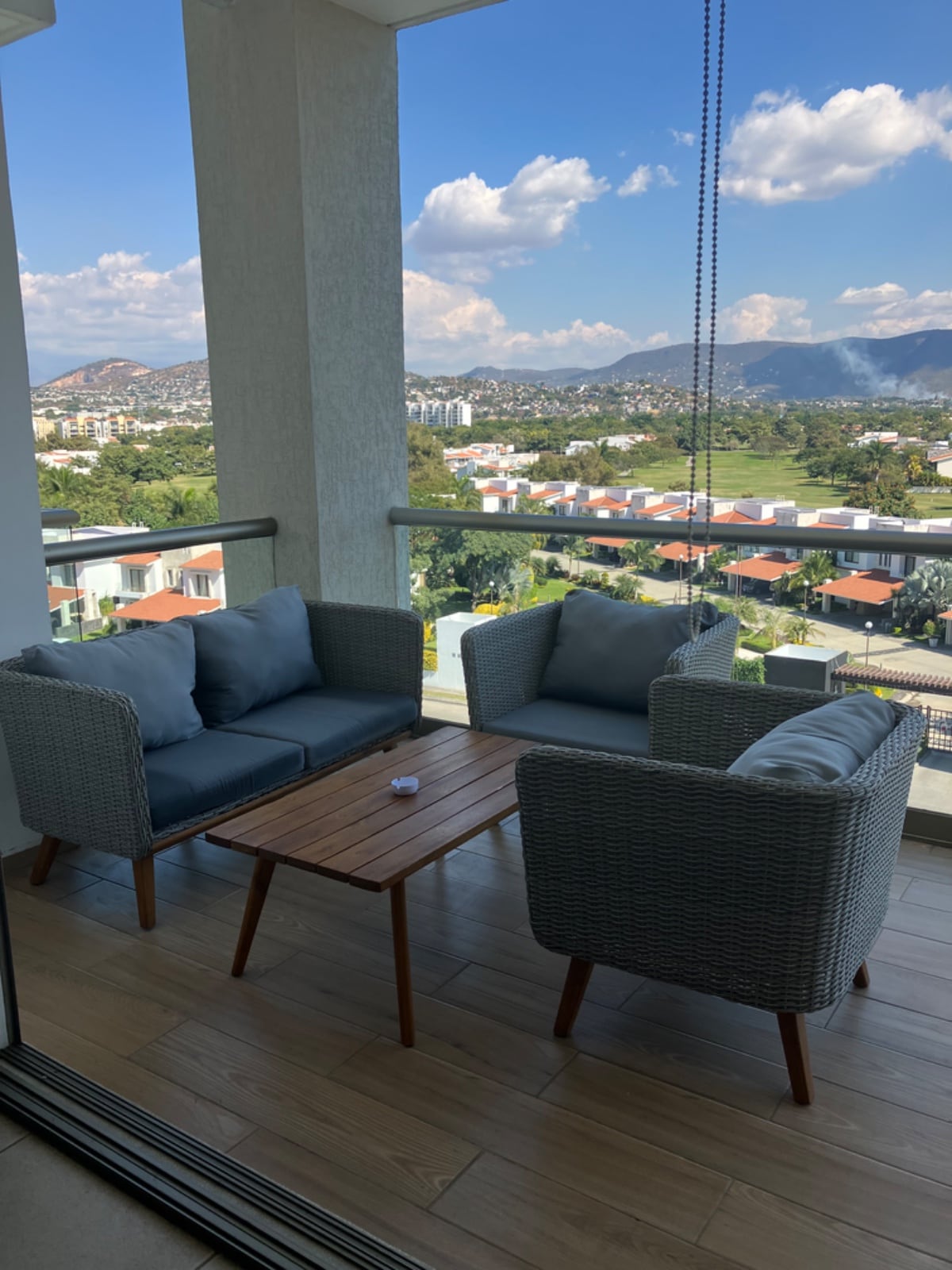 A balcony seating area is presented, featuring two woven chairs with plush cushions and a small wooden table. The backdrop includes a scenic view of rolling hills and golf course homes under a partly cloudy sky.