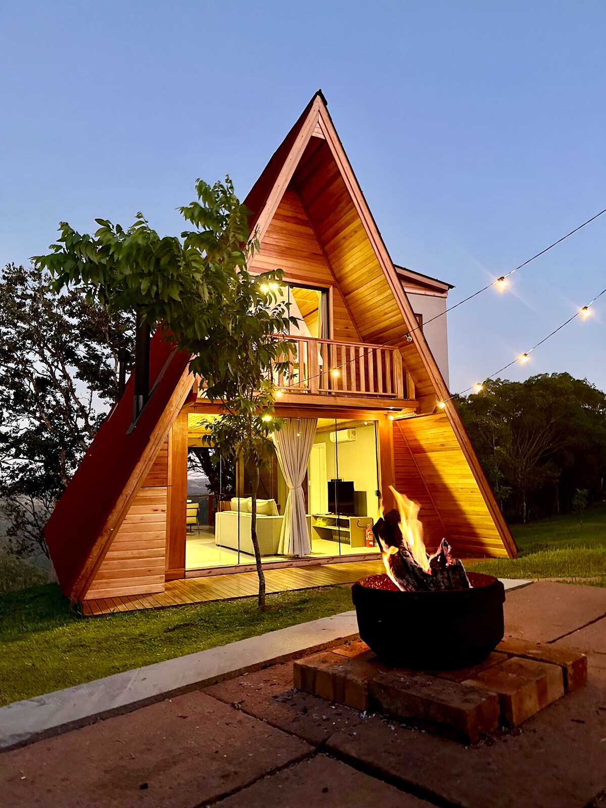An A-frame chalet is set in a grassy area, featuring large glass doors that open into a well-lit living space. A fire pit surrounded by stones is visible in the foreground, complementing outdoor string lights that add ambiance to the scene.