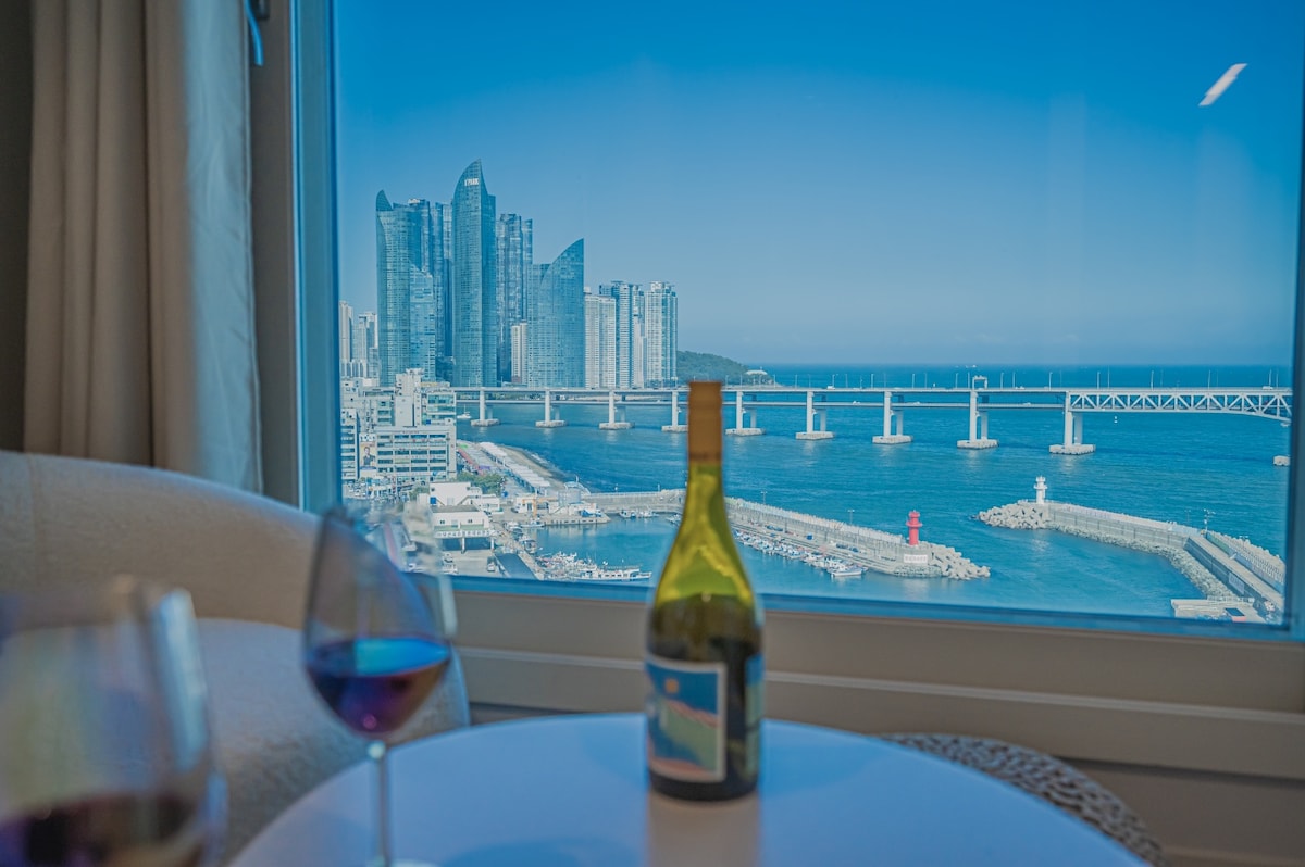 A clear view of the city skyline is visible through a large window, showcasing modern buildings against a blue sky. A bottle of wine and two glasses are placed on a table in the foreground, suggesting a leisurely atmosphere.