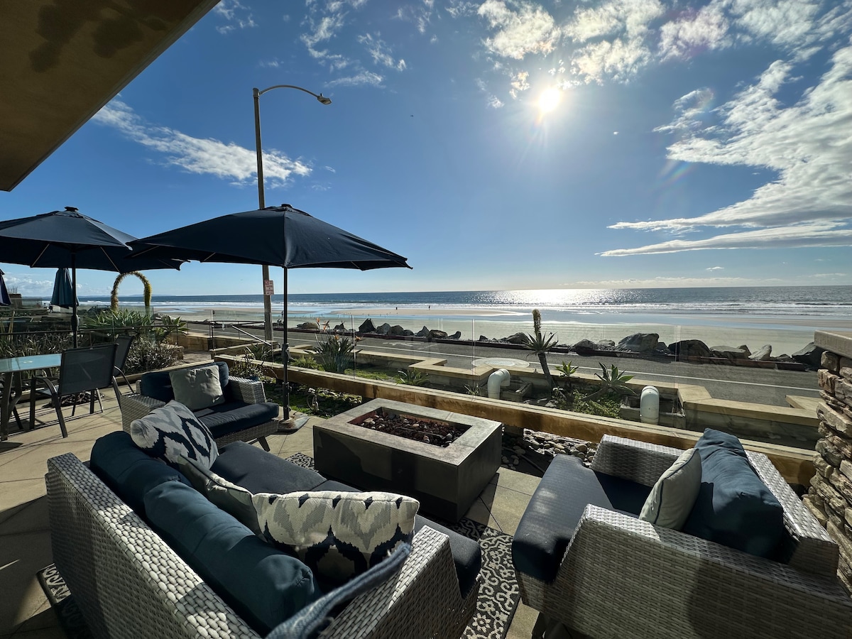 A stylish outdoor seating area is showcased, featuring modern furniture arranged around a gas fire pit. Umbrellas provide shade over the seating, while a view of the beach and ocean stretches beyond, with gentle waves lapping against the shore under a bright sky.