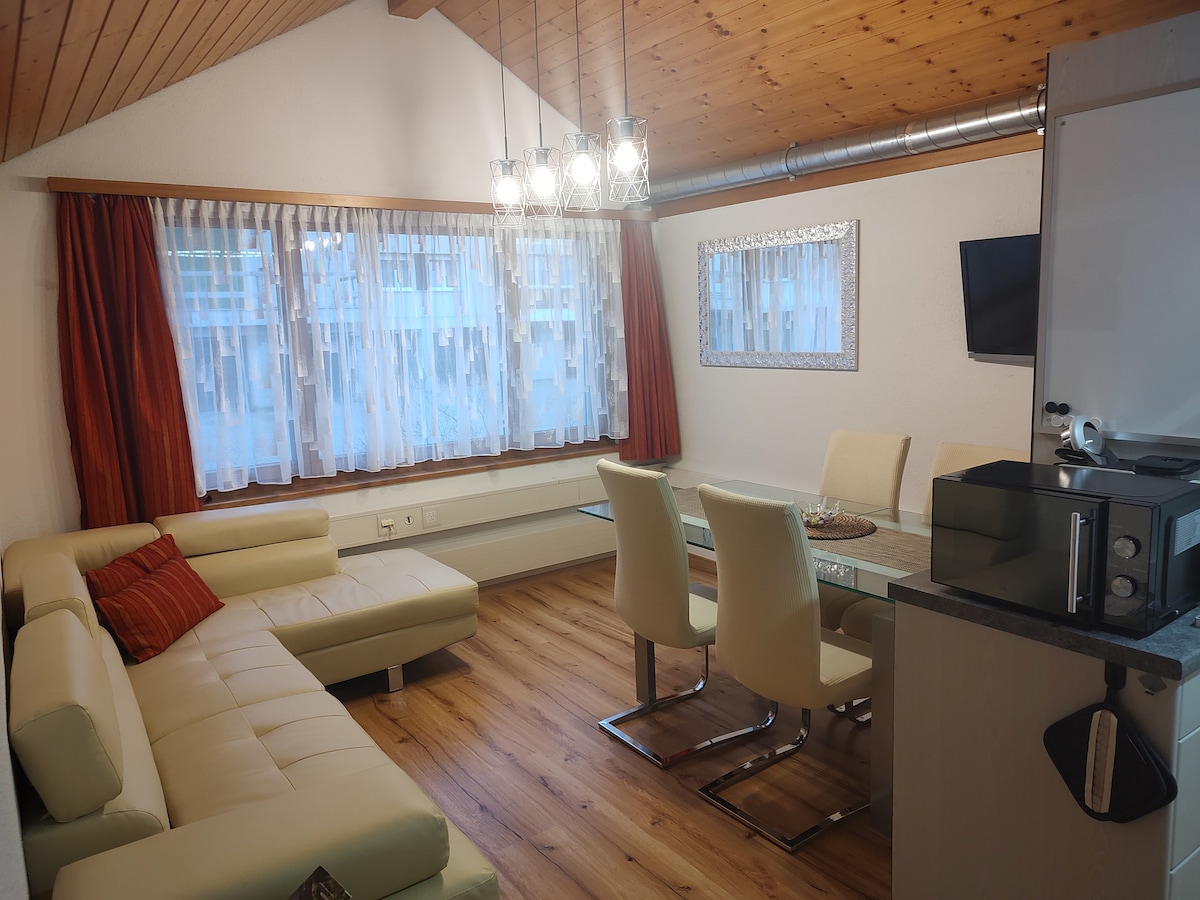 A spacious living area features a light-colored sectional sofa adorned with decorative pillows. A dining table with white chairs is positioned beside large windows fitted with sheer curtains. Wooden beams and a modern light fixture add character to the room.