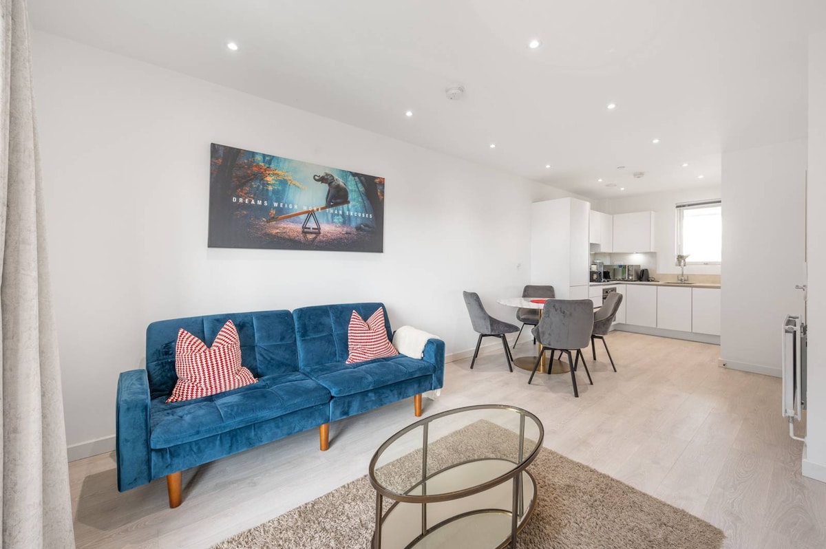 A modern living area features a blue sofa with red accent pillows and a round glass coffee table. The open plan design includes a dining table with four chairs and a well-equipped kitchen visible in the background. Recessed lighting enhances the bright interior.