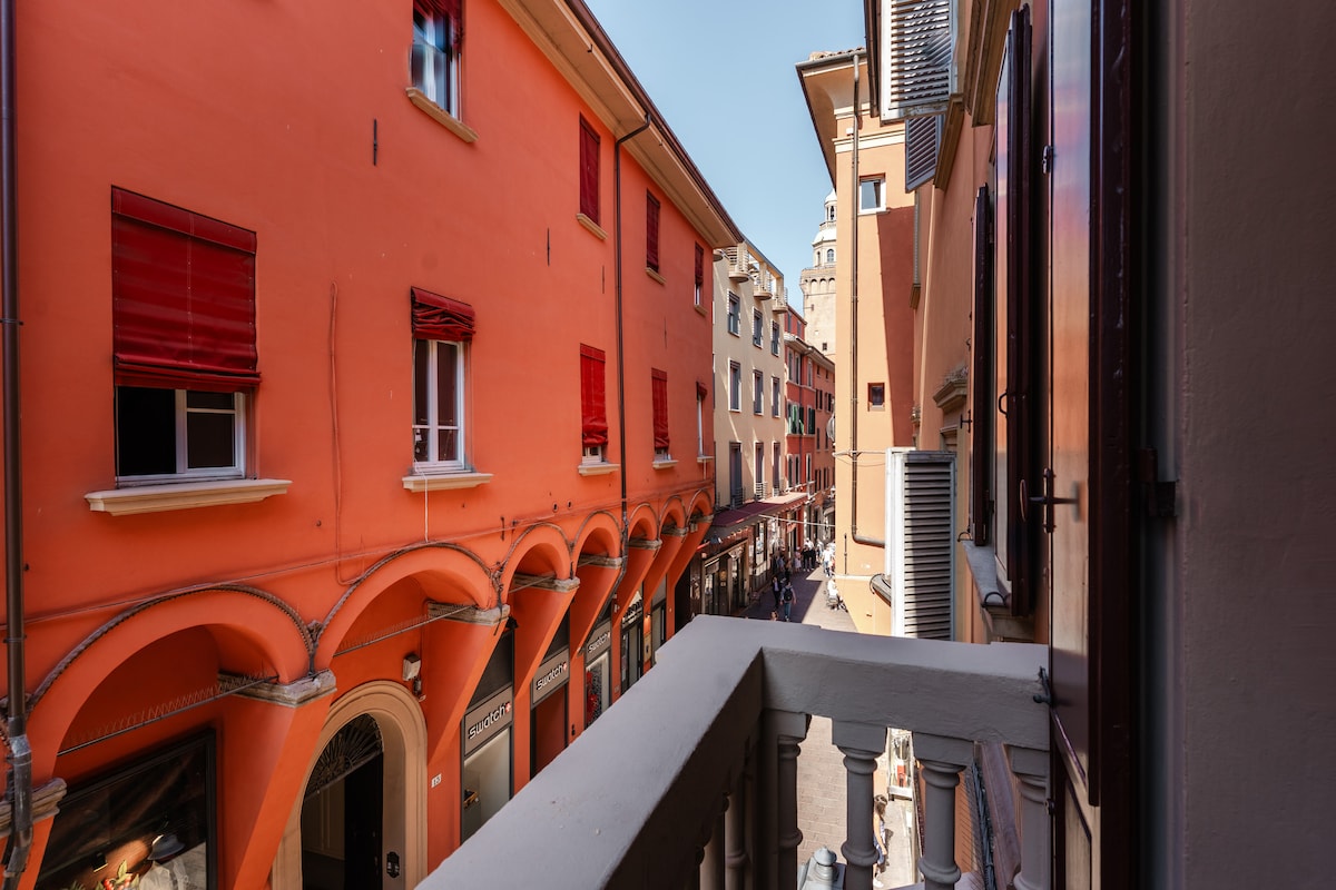 The view from the balcony showcases a charming street lined with orange-hued buildings. Red window shutters adorn the facades. Archways create a distinctive visual rhythm along the street, while pedestrians can be seen strolling beneath a clear blue sky.