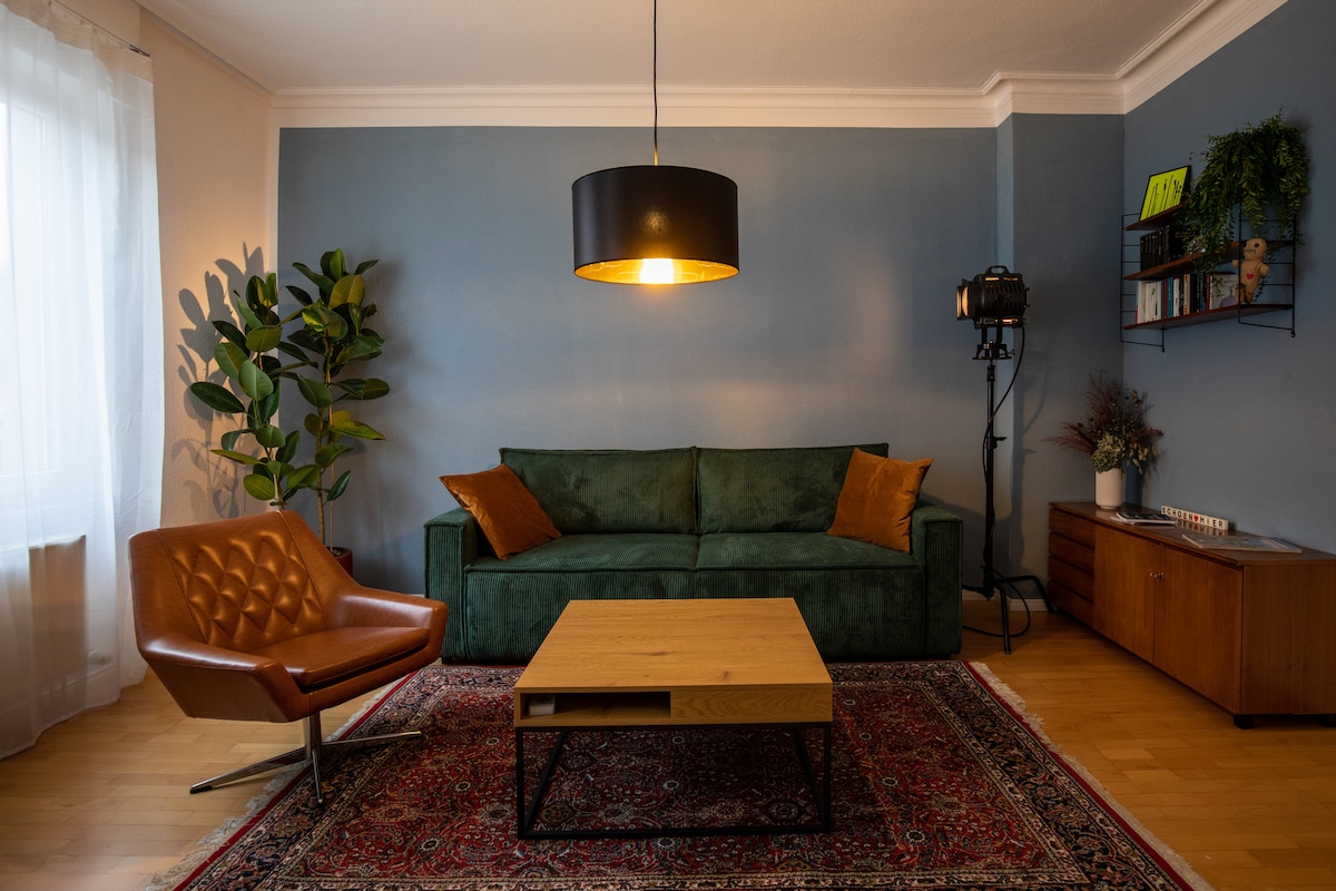 A spacious living area features a green sofa adorned with orange cushions, accompanied by a brown leather armchair. A wooden coffee table sits atop a decorative rug, while a black pendant light illuminates the room. A tall plant adds a touch of greenery.