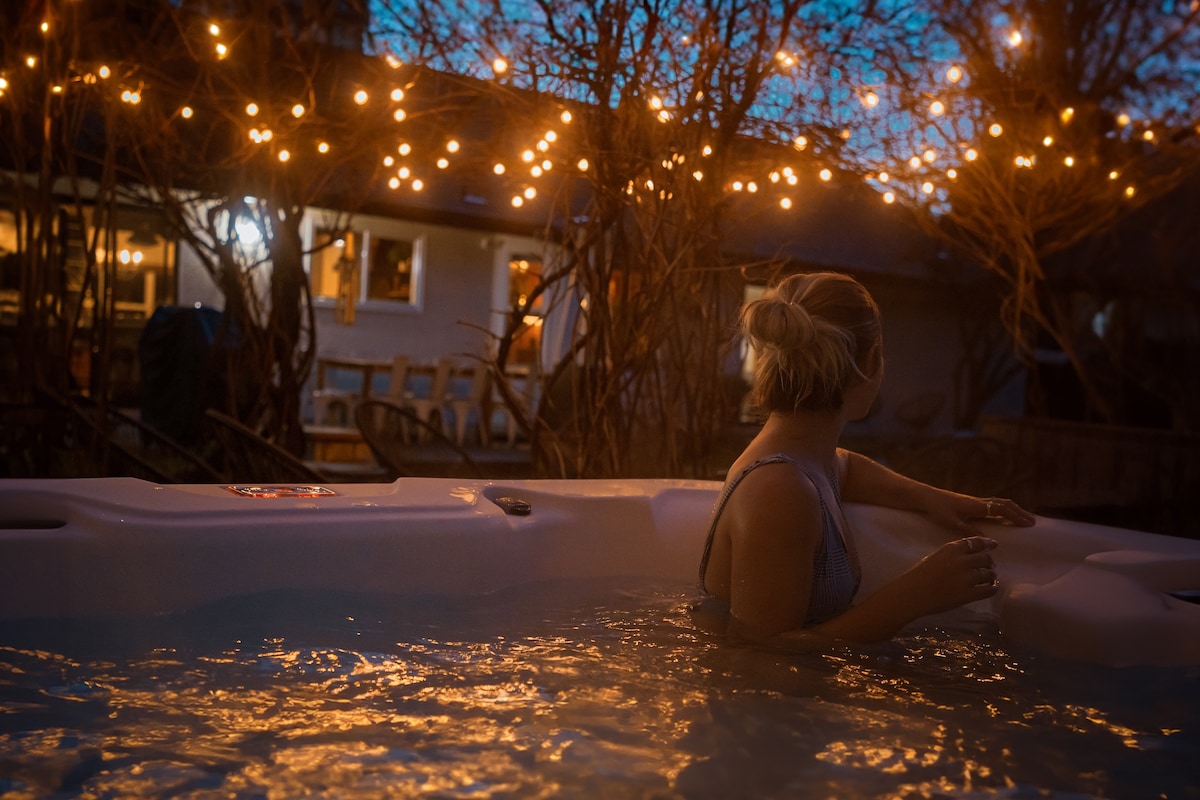 A relaxing hot tub is surrounded by softly glowing string lights, creating a warm ambiance in the evening. A guest can be seen enjoying the water, with the house's illuminated windows visible in the background, hinting at a welcoming atmosphere.