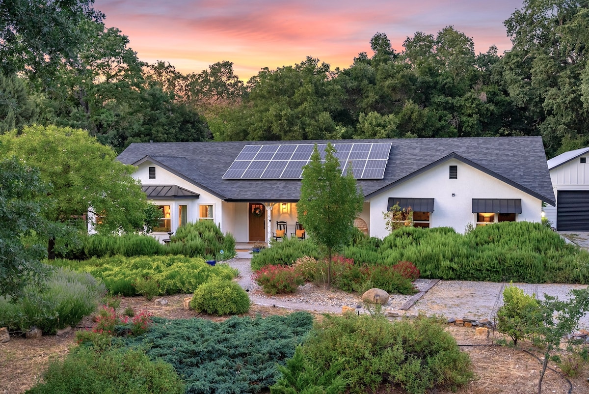 The exterior of the property is showcased during sunset, with solar panels on the roof and lush landscaping featuring a variety of plants. The welcoming entrance is accentuated by warm lights from the windows, surrounded by greenery and colorful flowering bushes.