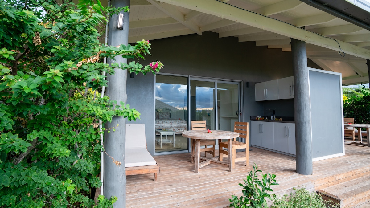 An outdoor deck area features a wooden table and chairs, with a kitchenette view through large glass doors. The space is surrounded by lush greenery, including climbing plants and flowering shrubs, creating a serene and inviting environment for relaxation.