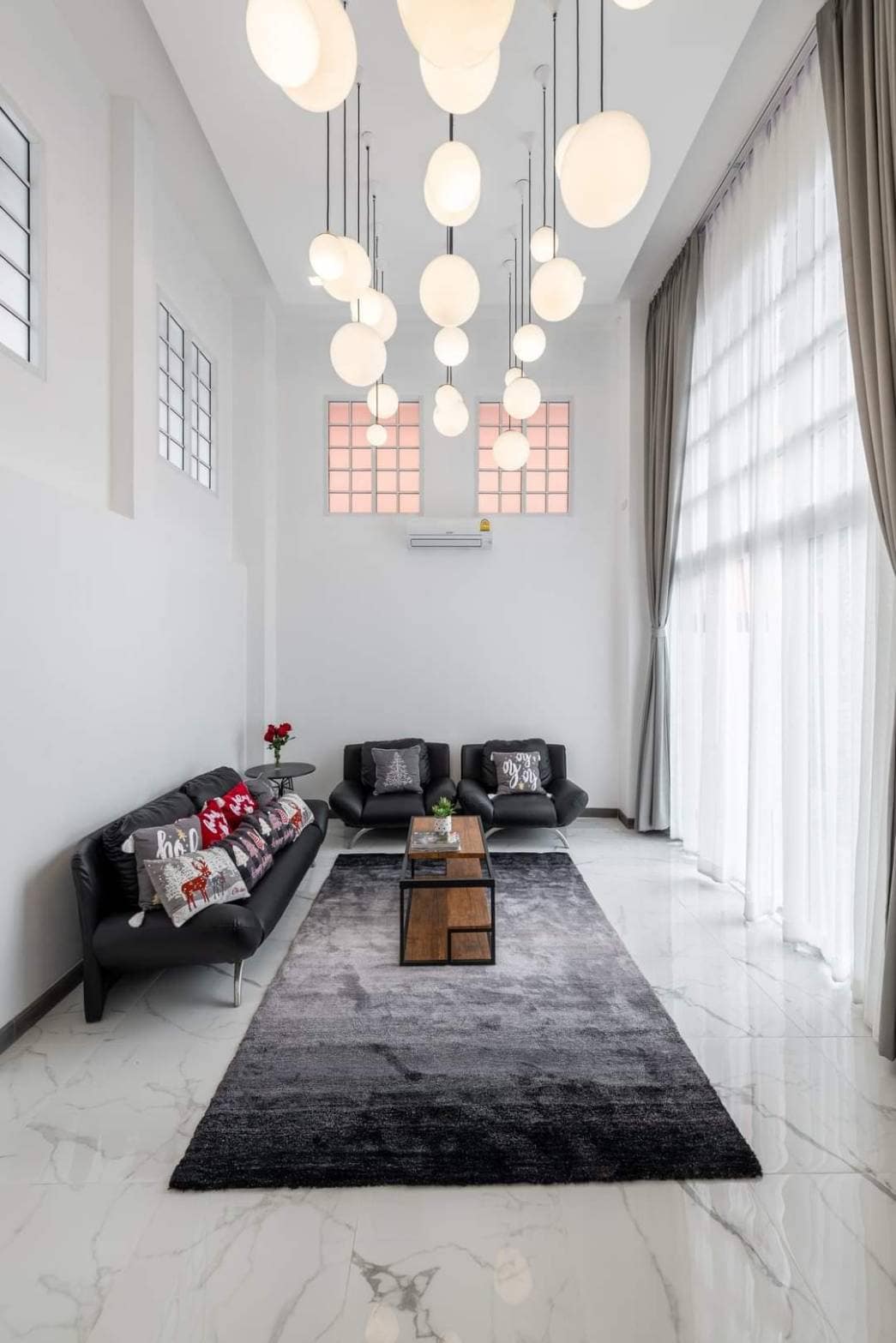 A modern living area is featured with high ceilings and large windows allowing natural light to fill the space. Comfortable black sofas with decorative pillows are arranged around a low wooden coffee table, set atop a plush gray rug, creating an inviting atmosphere.