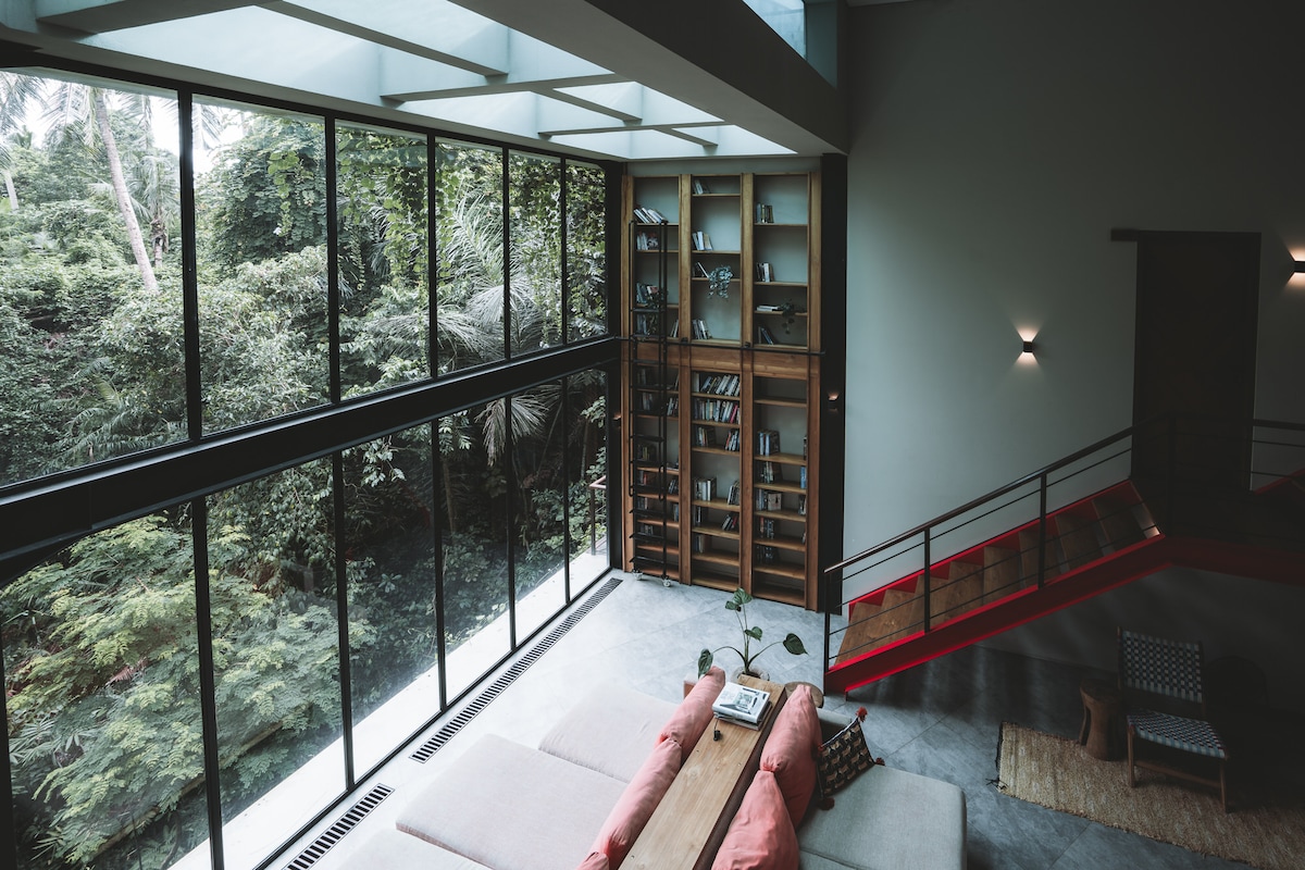 The lounge area is characterized by large floor-to-ceiling windows, framing views of lush greenery. A tall bookshelf stands against one wall, filled with books. An oversized, multi-facing couch is visible, offering a comfortable space for relaxation and viewing the surroundings.