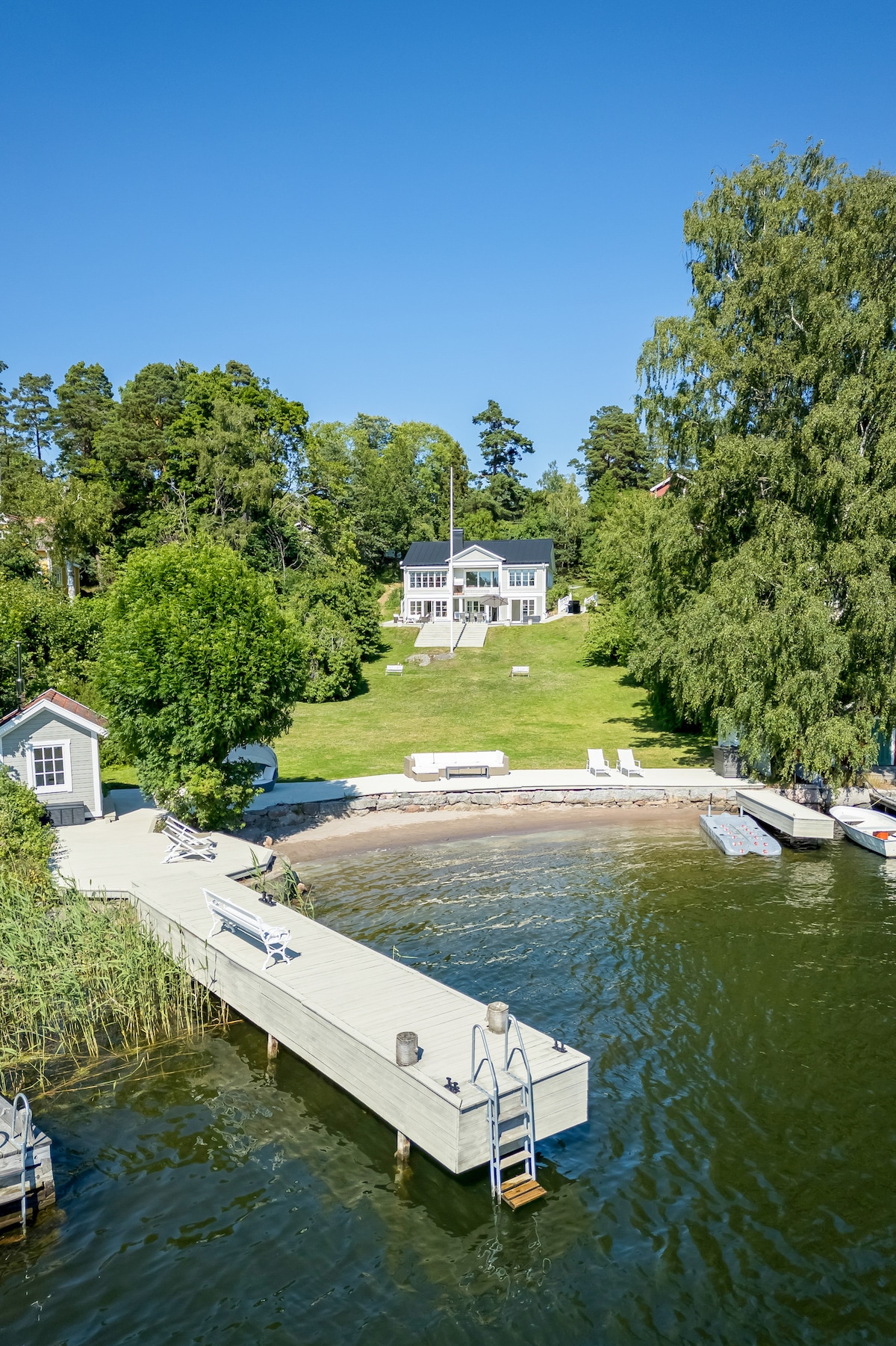 A spacious villa is visible from the water, surrounded by lush greenery. The property features a private dock with stairs leading into the water, alongside areas for seating and sunbathing. A well-maintained lawn complements the serene landscape.