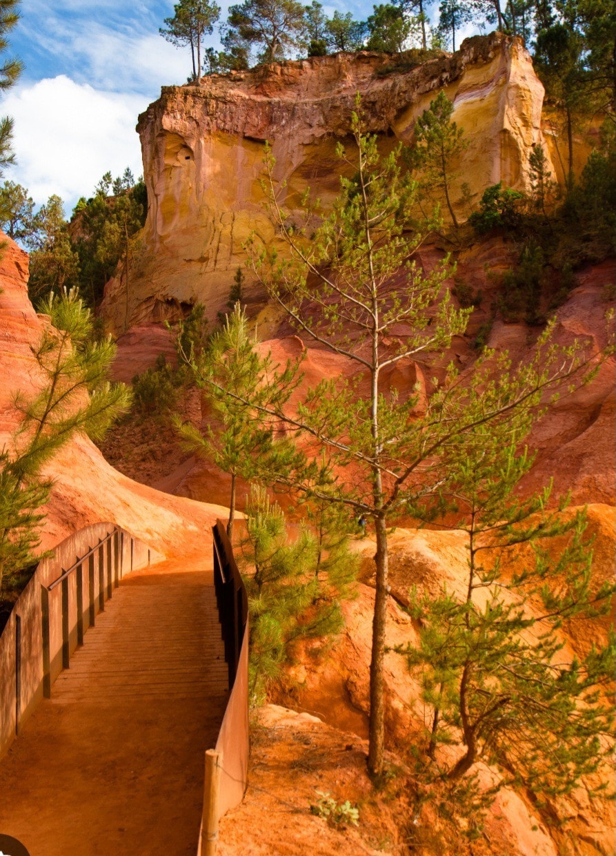 A wooden pathway winds through a naturally colored landscape, surrounded by towering cliffs in shades of red and yellow. Small pine trees are visible along the pathway, adding greenery to the striking scenery.