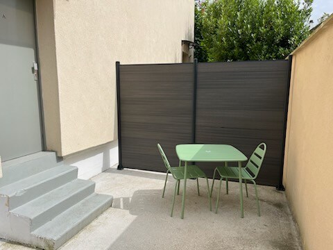 A small outdoor area features a green table surrounded by three matching chairs. A privacy screen is positioned behind the seating area. Steps lead up to an entrance, while shrubbery is visible in the background, providing a natural touch to the space.