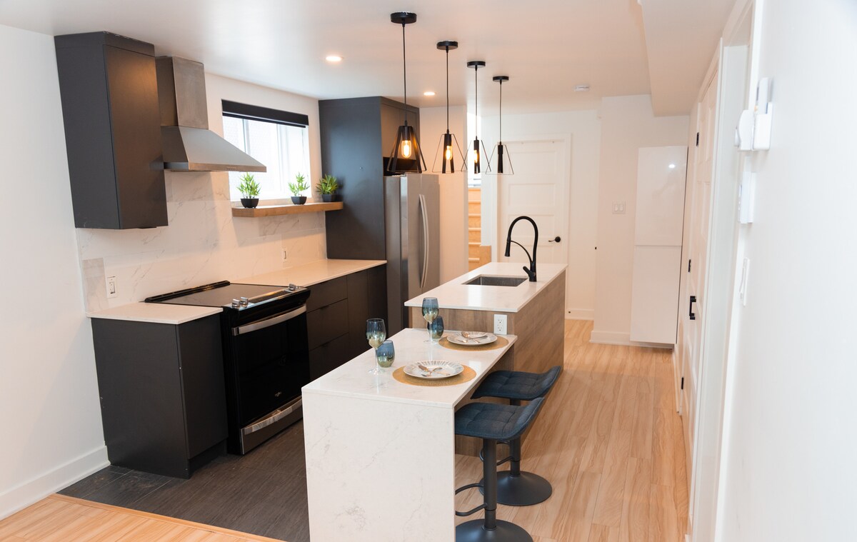 A modern kitchen is equipped with sleek black cabinetry and a spacious island. Stainless steel appliances include a range and refrigerator. Two blue upholstered chairs are positioned at the island, with decorative plates and glassware arranged on the countertop, complemented by greenery in small pots.