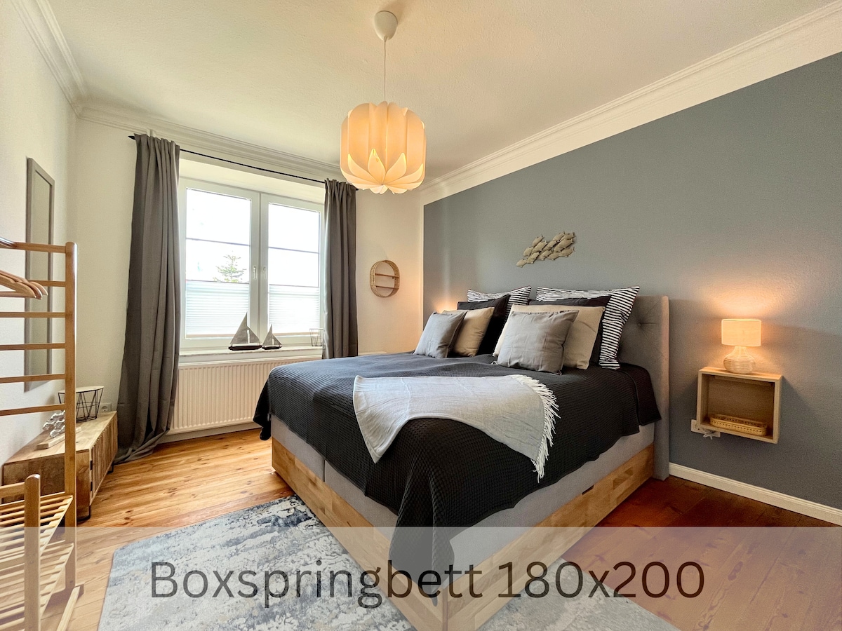 A spacious bedroom features a large boxspring bed with plush bedding and decorative accent pillows. Natural light enters through a window, complemented by soft gray curtains. Minimalist nightstands and a warm light fixture add to the calming atmosphere of the space.