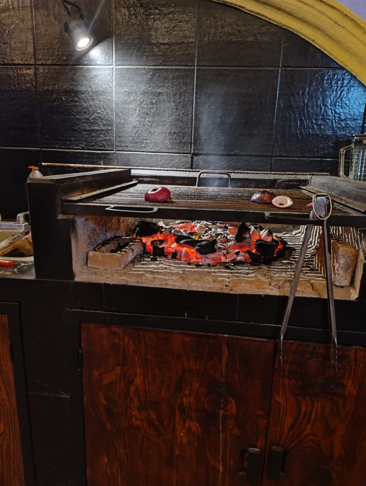 A traditional grilling station is situated against a sleek black wall. Charcoal glows warmly beneath grill grates, surrounded by various grilling utensils. The wooden cabinetry below contrasts with the modern elements, creating a functional setup for outdoor cooking.