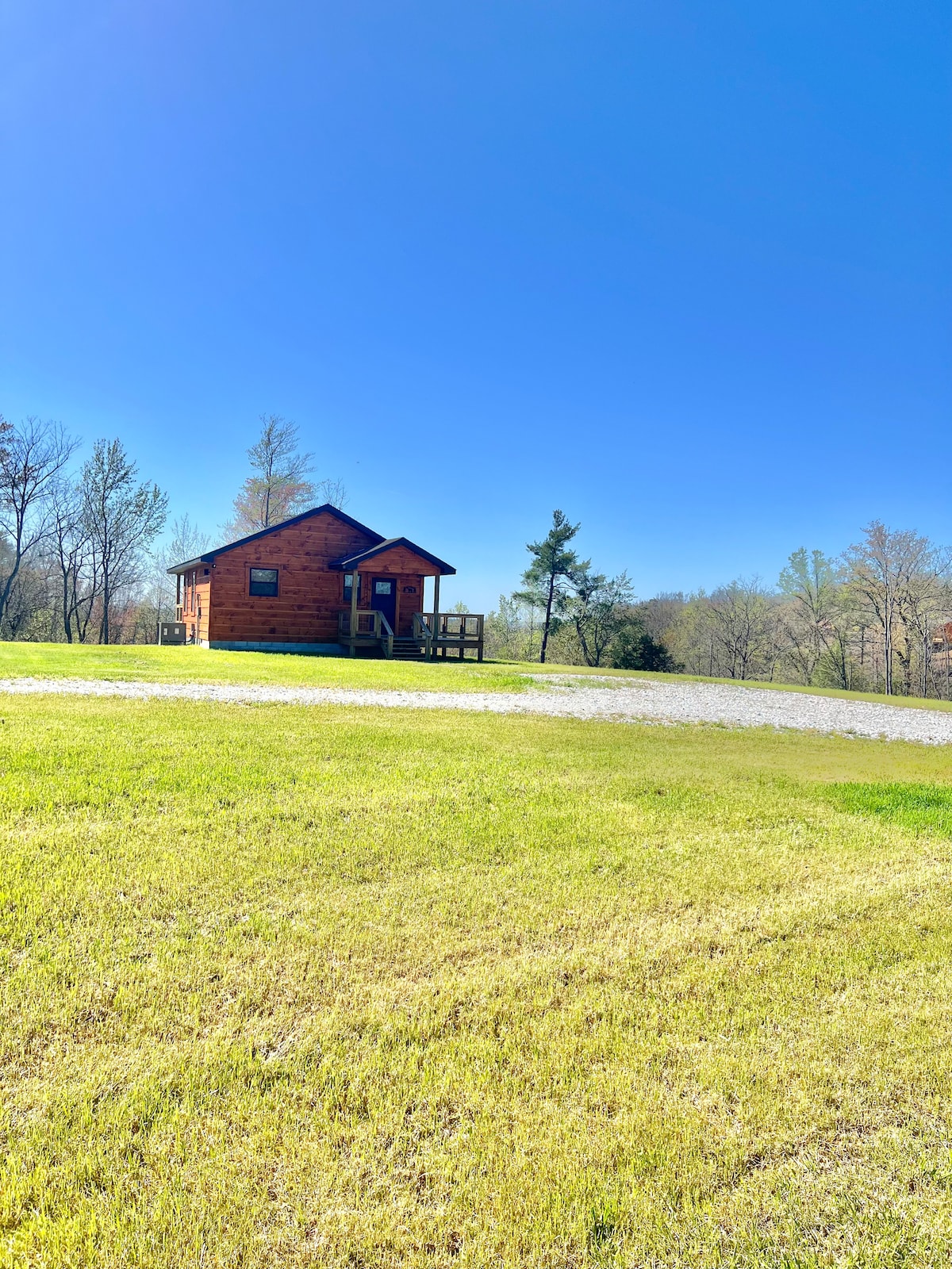 A charming cabin is nestled on a green grassy slope with a gravel driveway leading up to it. The clear blue sky above enhances the serene outdoor setting, surrounded by trees and distant landscapes.