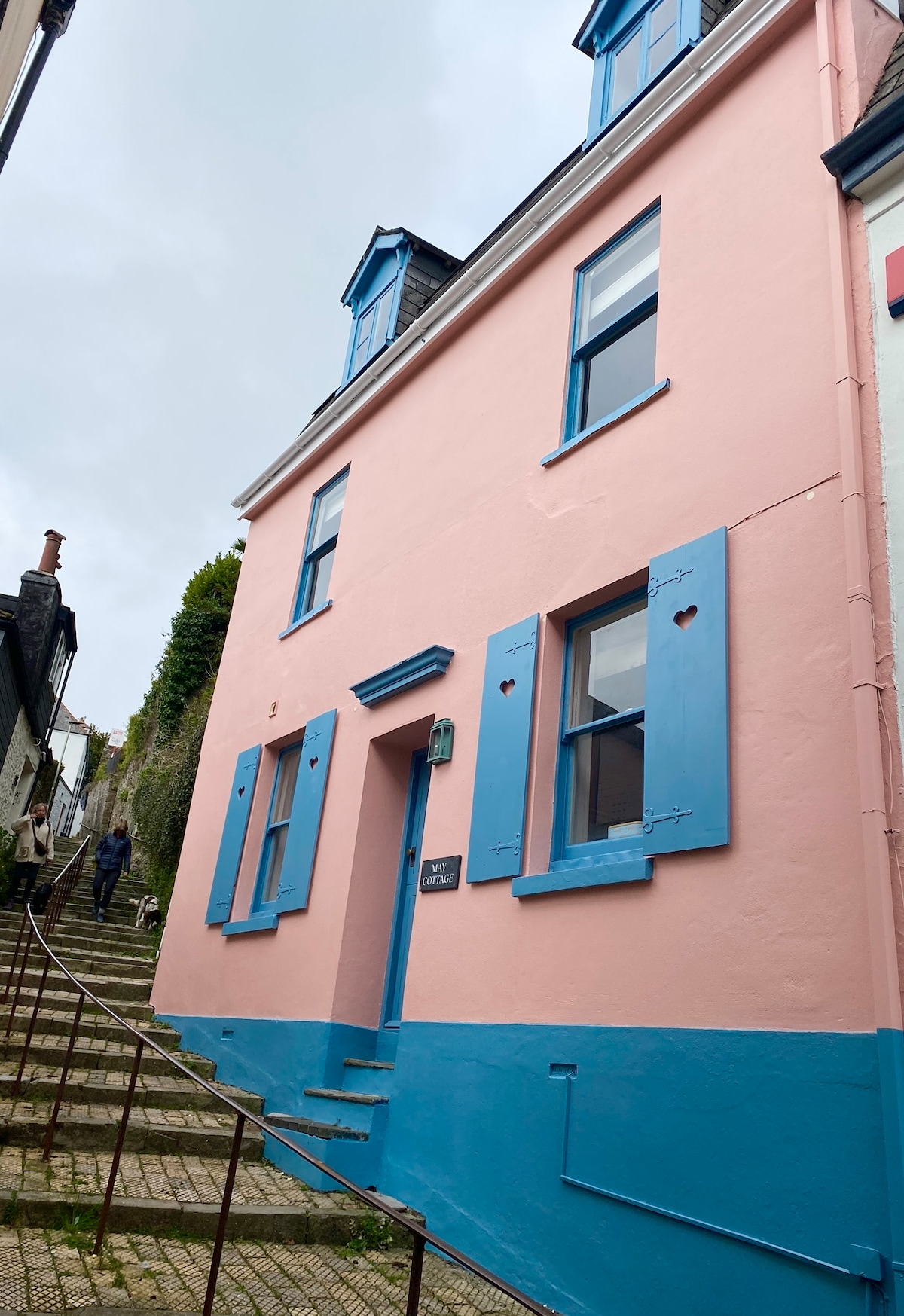 A charming cottage exterior features a pink façade with blue shutters and trim. The building is set on a narrow, steep path lined with stone steps. A person can be seen walking up the steps, emphasizing the elevated location of the property.