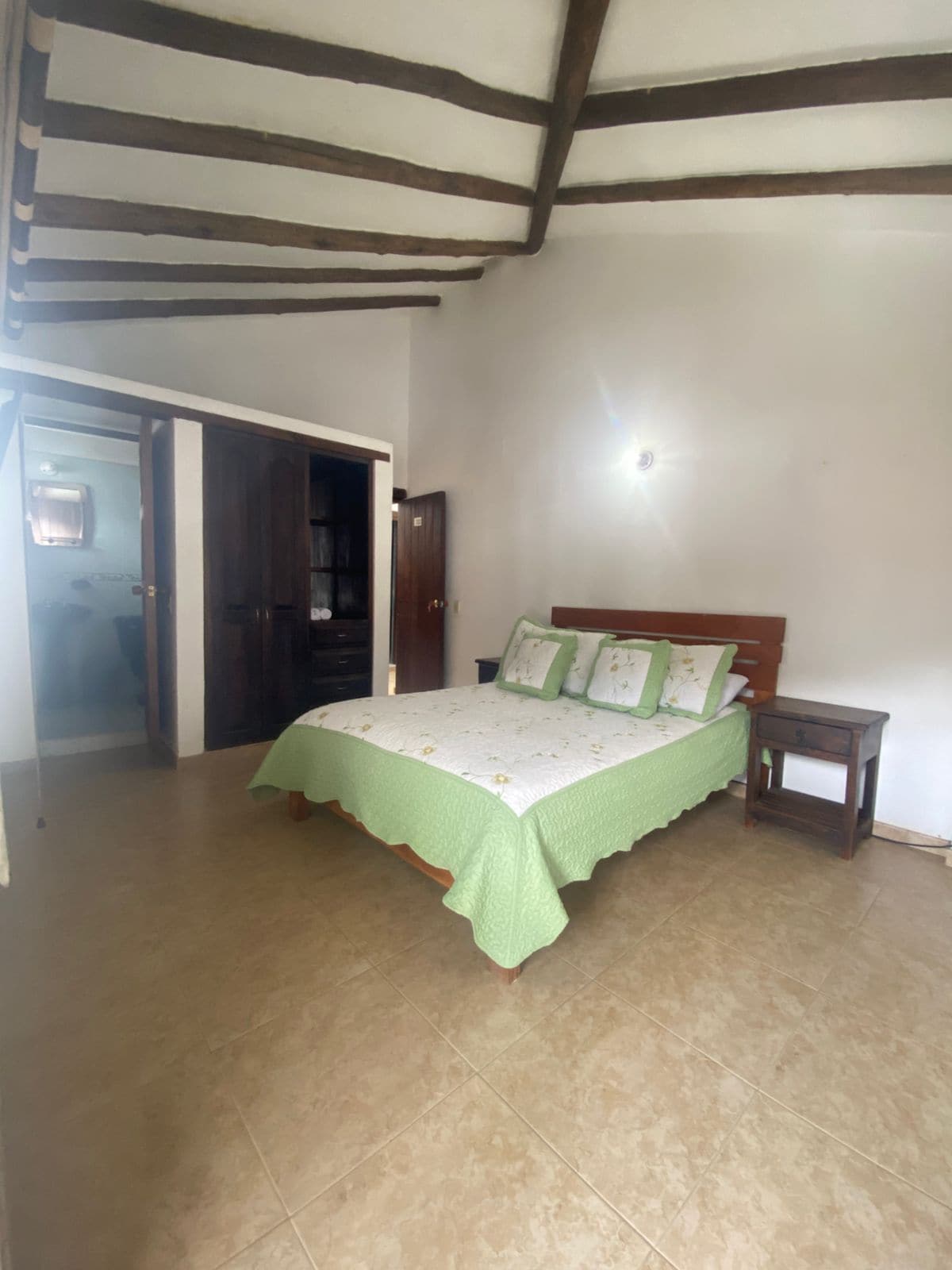 A double bed is dressed in a light green bedspread, accompanied by decorative pillows. A wooden nightstand sits beside the bed, and a closet is positioned against the wall. The room features bright wall colors and exposed wooden beams on the ceiling.