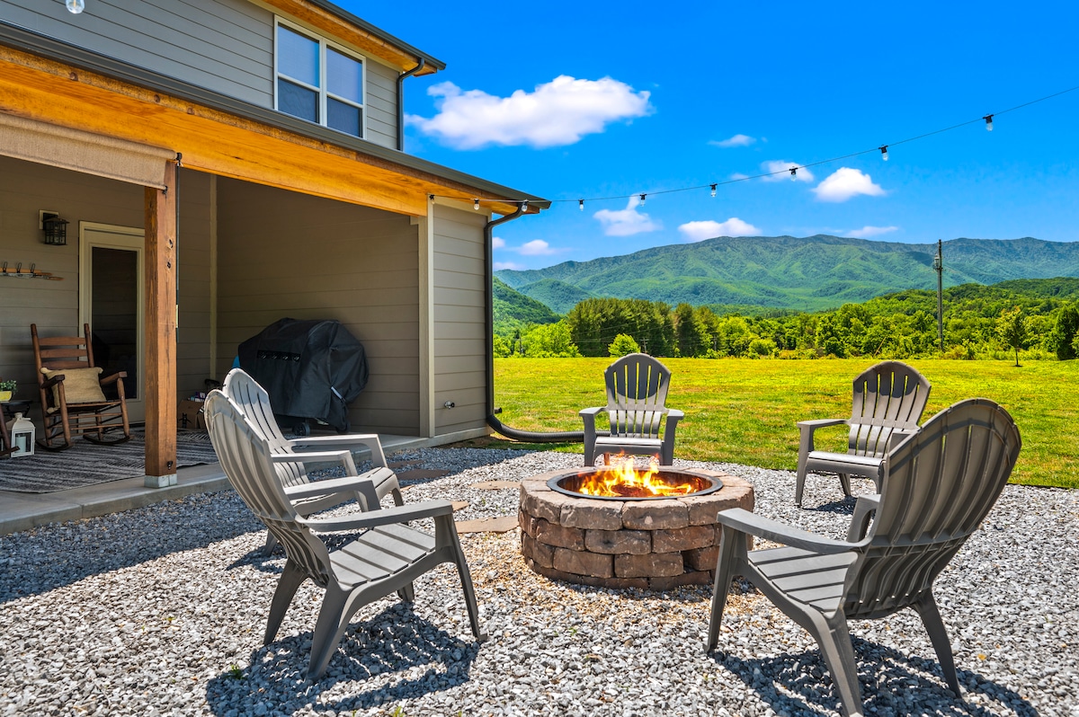 An outdoor fire pit area is surrounded by five gray chairs, situated on a gravel surface. A spacious backyard offers views of rolling green hills and mountains under a bright blue sky. The cabin features a covered patio with a grill in the background.
