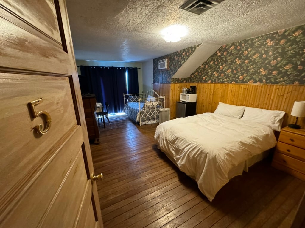 A warm room features a full bed with white bedding, positioned on polished wooden floors. A small work area with a microwave, refrigerator, and coffee maker is visible. Natural light enters through a window, partially covered by dark curtains. Floral wallpaper adds a charming touch.