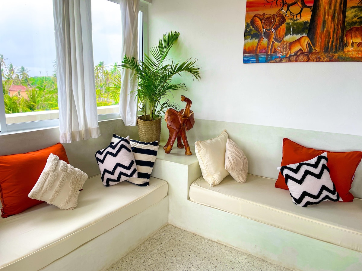 A cozy corner of the living area features built-in seating with soft cushions, including orange, striped, and geometric patterns. A small decorative wooden elephant and a potted plant add a touch of nature, while large windows allow natural light to fill the space.