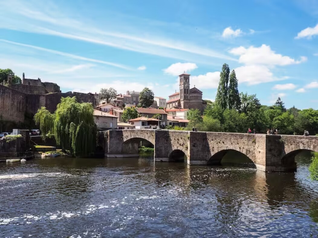A stone bridge arches over a calm river, connecting lush greenery with a charming village. Historical buildings and a church tower are framed by trees under a clear blue sky, while people can be seen walking along the bridge.
