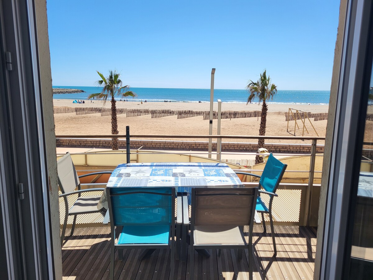 A dining table with six chairs is positioned on a balcony facing the beach. The expansive view includes a sandy shoreline and calm blue waters, framed by palm trees. A clear sky enhances the inviting outdoor space.