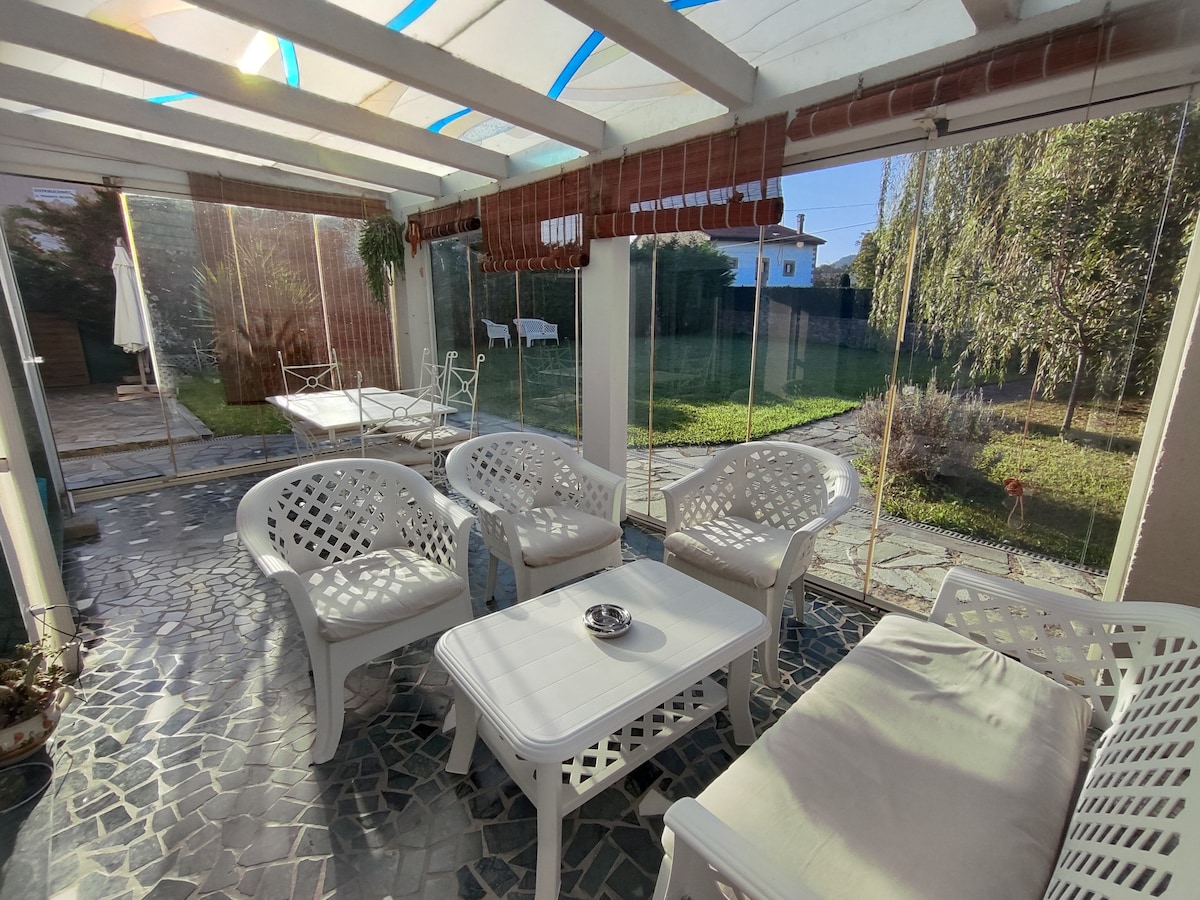 A bright sunroom features white wicker furniture arranged comfortably around a central table. Glass walls provide a clear view of the surrounding garden, allowing natural light to fill the space. A cozy seating area is complemented by a tranquil green backdrop.