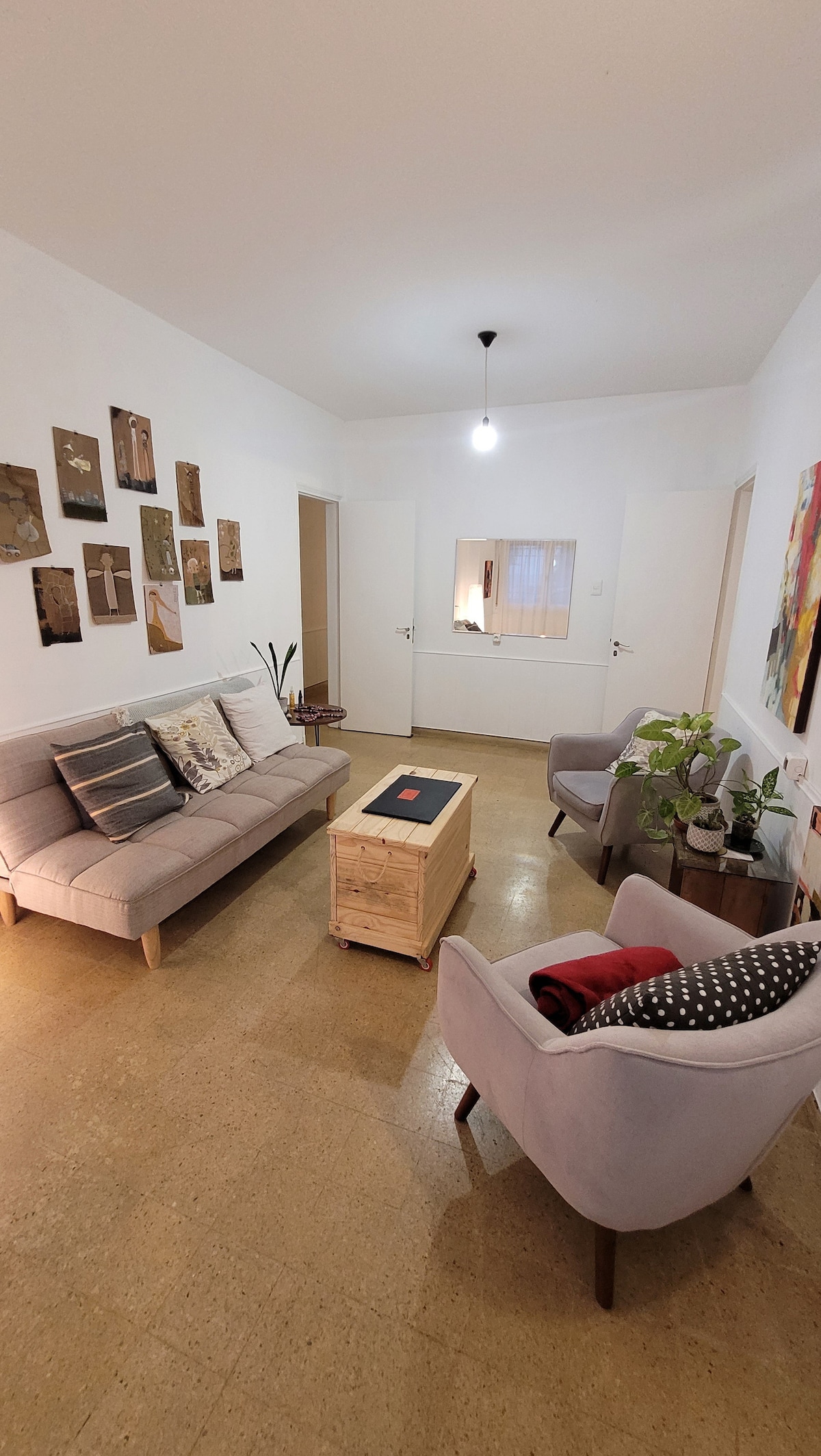 A bright living area contains a light-colored sofa and two armchairs, accompanied by a wooden coffee table at the center. The walls are adorned with various framed artworks, while a couple of plants add a touch of greenery to the space.