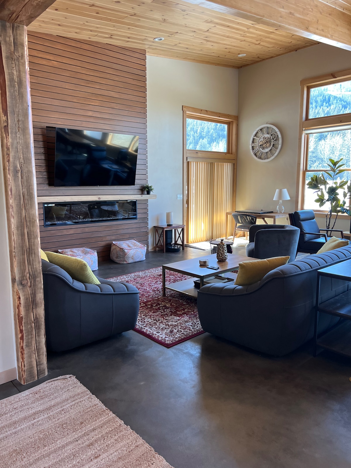 A spacious living area is highlighted by a large television mounted on a wooden accent wall. Comfortable seating arrangements include two dark sofas and a low coffee table, creating a cozy environment. Natural light fills the space through tall windows, enhancing the inviting atmosphere.