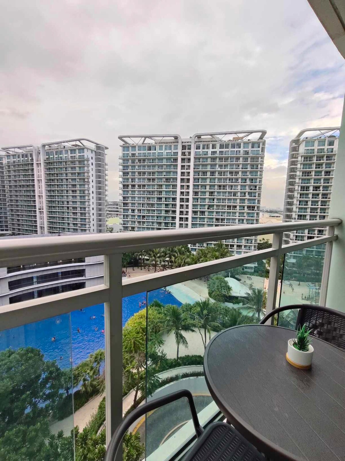 A balcony space features a small round table with two chairs overlooking a communal swimming pool and landscaped area below. Modern high-rise buildings rise in the background, framed by a subtle sky with soft clouds.