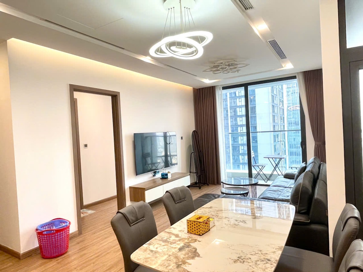 A modern living area is highlighted by a large window offering city skyline views. A comfortable sofa faces a flat-screen TV mounted on the wall. A dining table sits to the side, accompanied by stylish chairs. A laundry basket can be seen nearby, adding to the home's functionality.